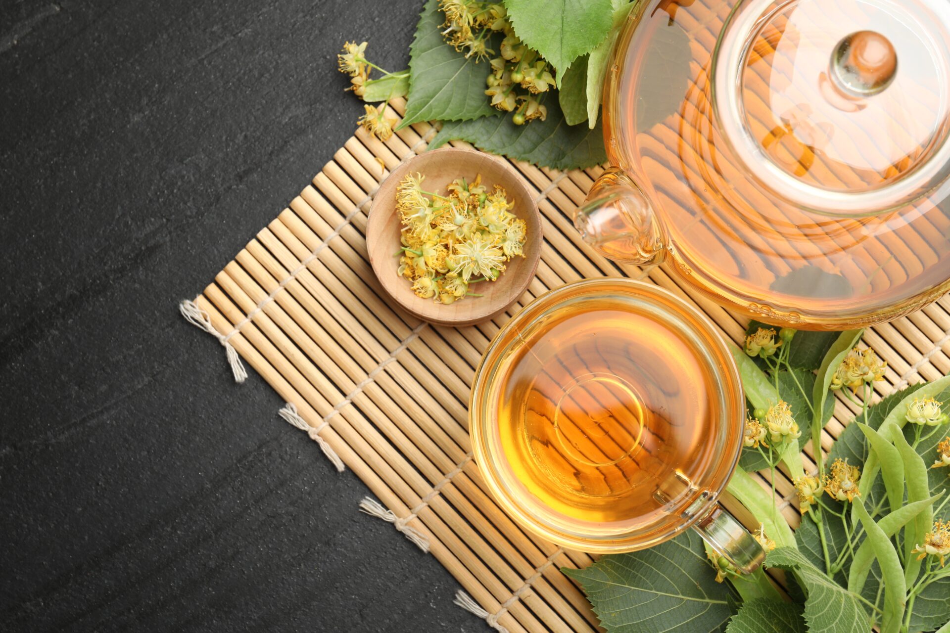 Tasty linden tea in cup, teapot, leaves and flowers on dark textured table, top view